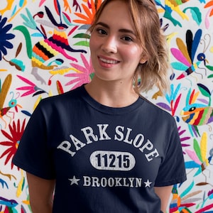 Op de afbeelding: Een marineblauw t-shirt met witte tekst die "Park Slope 11215 Brooklyn" zegt. Het shirt wordt gedragen door een persoon met lang bruin haar.