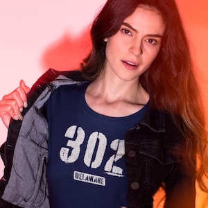 Könnte beinhalten: Eine Frau trägt ein marineblaues T-Shirt mit weißem Text, der "302 DELAWARE" lautet, und eine schwarze Jeansjacke. Sie steht vor einem rosa und orangenen Hintergrund.
