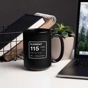 Puede incluir: Una taza de caf&eacute; negra con un gr&aacute;fico cuadrado blanco que dice "ELEMENT 115 (289) |Rn|5f&sup1;⁴6d&sup1;⁰7s&sup2;7p&sup3; Mc | Moscovium". La taza est&aacute; sobre una superficie blanca con un port&aacute;til, una planta suculenta y una cesta de alambre negra.