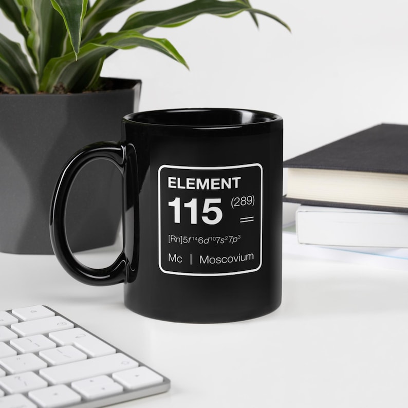 Puede incluir: Taza de cer&aacute;mica negra con un gr&aacute;fico cuadrado blanco. El gr&aacute;fico dice "ELEMENT 115 (289) = [Rn]5f&sup1;⁴6d&sup1;⁰7s&sup2;7p&sup3; Mc | Moscovium". La taza est&aacute; sobre una superficie blanca con un teclado, una planta y un libro en el fondo.