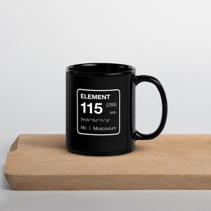 Puede incluir: Taza de cer&aacute;mica negra con un gr&aacute;fico cuadrado blanco que dice "ELEMENT 115 (289)" y el s&iacute;mbolo qu&iacute;mico del Moscovio. La taza est&aacute; sobre una superficie de madera.