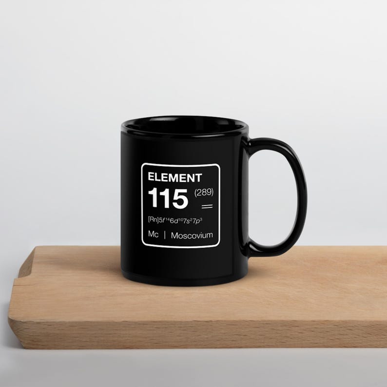 Puede incluir: Taza de cer&aacute;mica negra con un gr&aacute;fico cuadrado blanco que dice "ELEMENT 115 (289)" y el s&iacute;mbolo qu&iacute;mico del Moscovio. La taza est&aacute; sobre una superficie de madera.