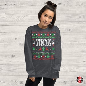 May include: A dark grey Christmas jumper with a white design. The design features the word "MOZ" in a retro font, two stylised faces, a reindeer, a Christmas tree, and the text "There is a light that never goes out".