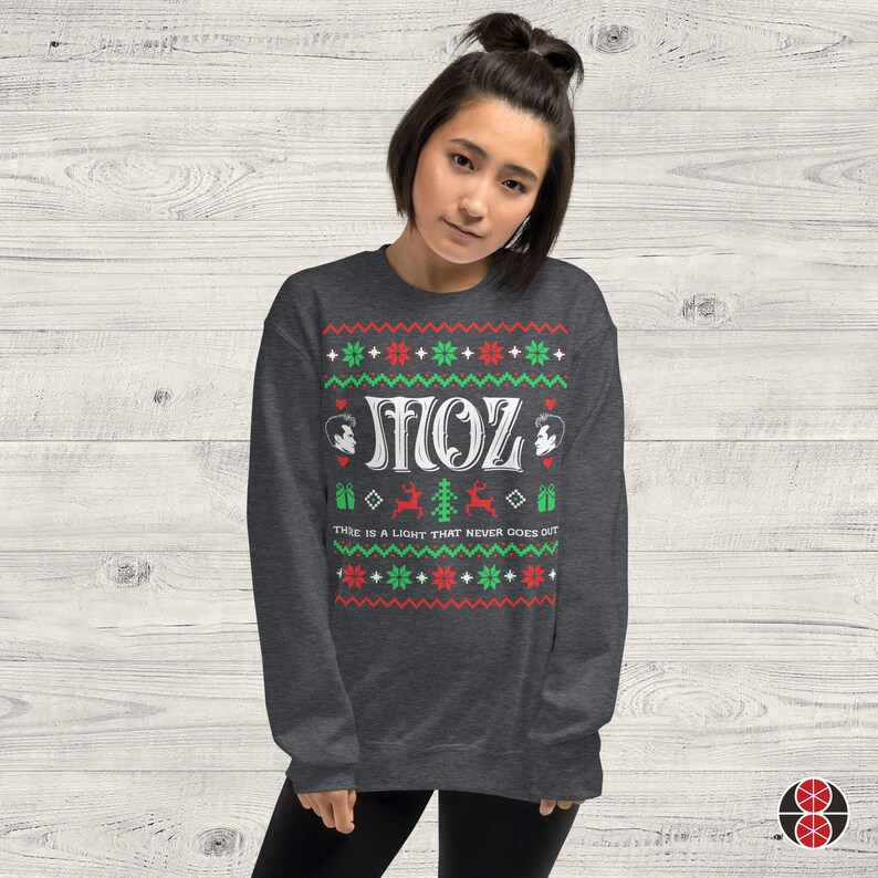 May include: A dark grey Christmas jumper with a white design. The design features the word "MOZ" in a retro font, two stylised faces, a reindeer, a Christmas tree, and the text "There is a light that never goes out".