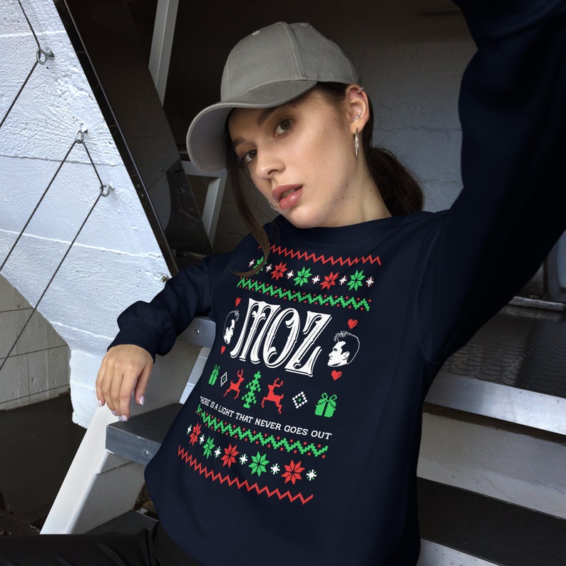 May include: A navy blue Christmas jumper with a white design featuring the text "Moz" and a reindeer. The jumper has a red and green pattern with snowflakes and hearts. The text "There is a light that never goes out" is printed below the reindeer.
