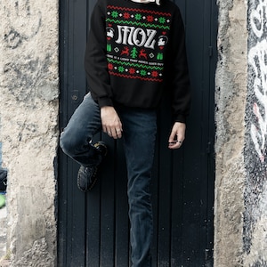 May include: A black Christmas jumper with a green and red pattern. The jumper has the text "MOZ" in white letters and the phrase "There is a light that never goes out" in smaller white letters.