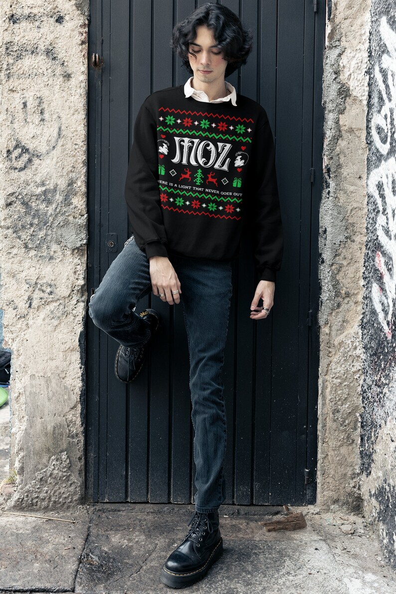 May include: A black Christmas jumper with a green and red pattern. The jumper has the text "MOZ" in white letters and the phrase "There is a light that never goes out" in smaller white letters.