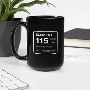 Puede incluir: Taza de cer&aacute;mica negra con el texto "ELEMENT 115 (289) = [Rn]5f&sup1;⁴6d&sup1;⁰7s&sup2;7p&sup3; Mc | Moscovium" en un rect&aacute;ngulo blanco. La taza tiene un asa grande y est&aacute; sobre una superficie blanca.