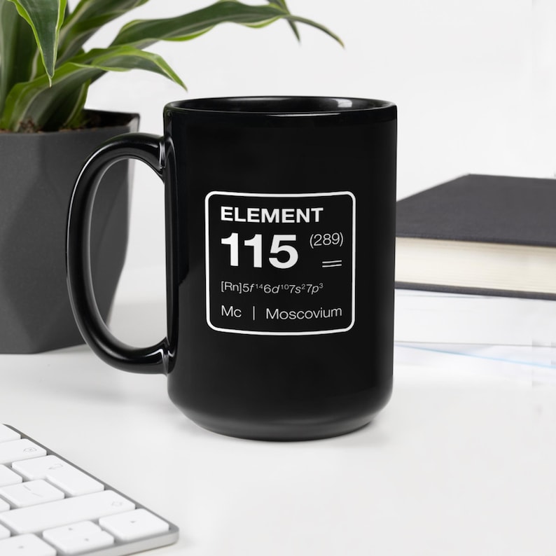 Puede incluir: Taza de cer&aacute;mica negra con el texto "ELEMENT 115 (289) = [Rn]5f&sup1;⁴6d&sup1;⁰7s&sup2;7p&sup3; Mc | Moscovium" en un rect&aacute;ngulo blanco. La taza tiene un asa grande y est&aacute; sobre una superficie blanca.