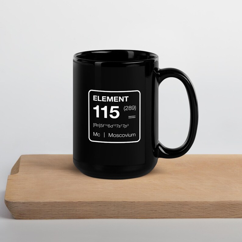 Puede incluir: Una taza de cer&aacute;mica negra con un gr&aacute;fico cuadrado blanco que muestra la palabra "ELEMENT", el n&uacute;mero "115" y el s&iacute;mbolo qu&iacute;mico del Moscovio. La taza est&aacute; sobre una superficie de madera.