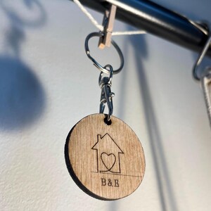 Op de afbeelding: Een ronde houten sleutelhanger met een huis- en hartontwerp, en de letters "B&B" eronder gegraveerd. De sleutelhanger is bevestigd aan een zilveren karabijnhaak en ring. De achtergrond is een lichtgrijze muur.