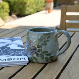 May include: A ceramic mug with a floral design in shades of blue, green, and white. The mug is sitting on a wooden table next to a magazine with the text "Rue Cambon" on the cover.