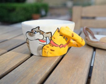 Puede incluir: Una taza de cerámica blanca con un diseño pintado a mano de dos gatos. Un gato es amarillo con manchas negras y el otro es gris con rayas marrones. El gato amarillo tiene una pajarita roja.