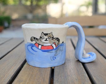 Puede incluir: Una taza de cerámica azul y blanca con un gato que lleva un salvavidas y nada en el océano. La taza tiene un asa ondulada única.