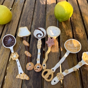 May include: Six ceramic spoons with unique bakery-themed handles and bowls. The designs include a dark chocolate spoon, a baguette-wrapped handle, a swirl pattern, a gingerbread man accent, a waffle-textured bowl, and a cupcake-shaped spoon.