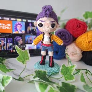 Puede incluir: Muñeca de ganchillo hecha a mano con un moño morado, que lleva una chaqueta amarilla, pantalones cortos azules y botas negras. La muñeca está sobre una base de ganchillo azul. Una pantalla de ordenador está al fondo.