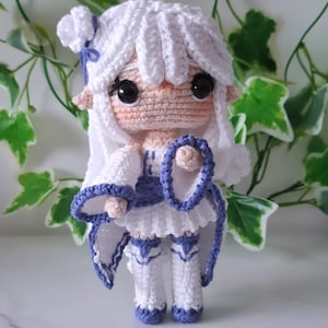 May include: A handmade crochet doll with white hair and a white dress with blue accents. The doll has large black eyes and pointed ears. It is holding a blue ring-shaped accessory. The doll is approximately 6 inches tall.