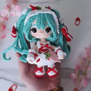 May include: A handmade crochet doll with turquoise hair, wearing a white dress with red accents and red shoes. The doll has large black eyes and is decorated with red bows. The doll is a unique, handcrafted item.