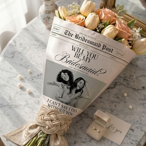 May include: A bouquet of peach roses and cream tulips wrapped in paper printed with The Bridesmaid Post, Will You Be My Bridesmaid?, and a black and white photo of two people. The stems are tied with a decorative gold braided ribbon and pearls.