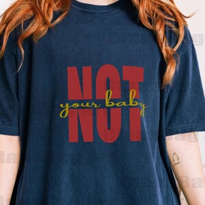 Puede incluir: Una camiseta azul marino con las palabras "NOT your baby NOT" en letras rojas y "your baby" en cursiva amarilla. La camiseta tiene un corte holgado y una etiqueta "TRUE NAVY".