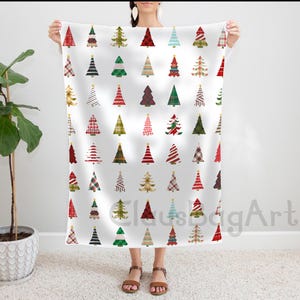 Puede incluir: Una manta blanca con un patrón repetido de árboles de Navidad en varios colores y diseños. Los árboles incluyen patrones rojos, verdes, dorados y a cuadros. Cada árbol tiene una estrella dorada en la parte superior. La manta es sostenida por una persona.