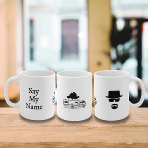 Puede incluir: Tres tazas de cerámica blanca con gráficos y texto en negro. Una taza tiene el texto "Say My Name", otra presenta una autocaravana con una nube, y la tercera muestra la silueta de un hombre con sombrero y gafas de sol.