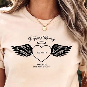 May include: A beige t-shirt with a black graphic of a heart with wings and a halo. The text "In Loving Memory" is above the heart and the text "ADD PHOTO" is inside the heart. The text "NAME HERE 01.05.1954 - 12.08.2023" is below the heart.