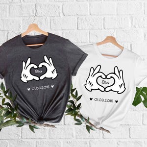 May include: Two t-shirts, one dark gray and one white, each featuring a graphic of white cartoon hands forming a heart shape. The word "Since" is inside the heart, with the date "01.03.2015" below. Both shirts are hanging on wooden hangers.