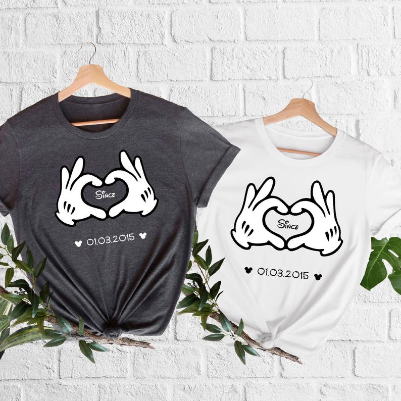 May include: Two t-shirts, one dark gray and one white, each featuring a graphic of white cartoon hands forming a heart shape. The word "Since" is inside the heart, with the date "01.03.2015" below. Both shirts are hanging on wooden hangers.