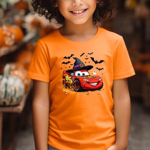 Puede incluir: Una camiseta naranja con un gr&aacute;fico de un coche de carreras rojo con el n&uacute;mero 95, que lleva un sombrero de bruja negro. El dise&ntilde;o incluye siluetas negras de murci&eacute;lagos voladores, un peque&ntilde;o fantasma blanco y trozos de caramelo de ma&iacute;z.