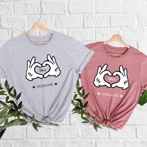 May include: Two t-shirts, one gray and one dusty rose, each featuring a heart made of white gloved hands. The word "Since" is inside the heart, with the date "01.03.2015" below. Both shirts are on wooden hangers.