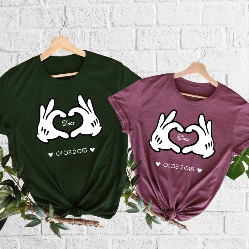 May include: Two t-shirts, one dark green and one burgundy, displayed on wooden hangers. Each shirt features a white graphic of gloved hands forming a heart, with the word "Since" inside and the date "01.03.2015" below.