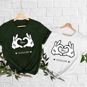 May include: Two t-shirts, one dark green and one white, each featuring a heart made of cartoon hands. The word "Since" is inside the heart, with the date "01.03.2015" below. Both shirts are hanging on wooden hangers.