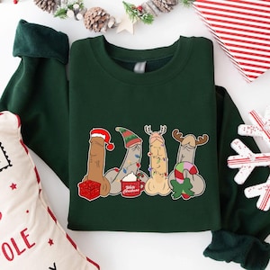May include: Dark green sweatshirt featuring a humorous Christmas design. The graphic depicts four phallic figures, each adorned with festive holiday elements like a Santa hat, elf hat, reindeer antlers, and candy cane. A mug with the words "White Christmas" is also included.