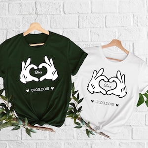 May include: Two t-shirts, one dark green and one white, hanging on wooden hangers. Each shirt features a graphic of cartoon hands forming a heart, with the word "Since" inside the heart and the date "01.03.2015" below.
