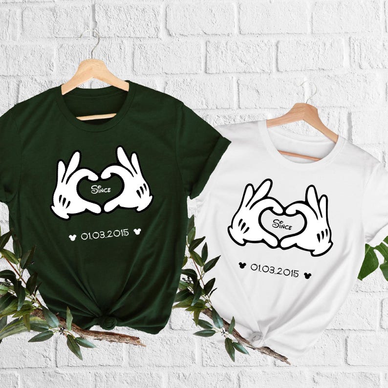 May include: Two t-shirts, one dark green and one white, hanging on wooden hangers. Each shirt features a graphic of cartoon hands forming a heart, with the word "Since" inside the heart and the date "01.03.2015" below.
