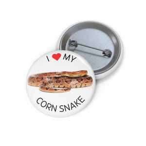 Puede incluir: Un botón blanco con un corazón rojo y el texto "Me encanta mi serpiente de maíz" en negro. Una serpiente de maíz enroscada se muestra debajo del texto.