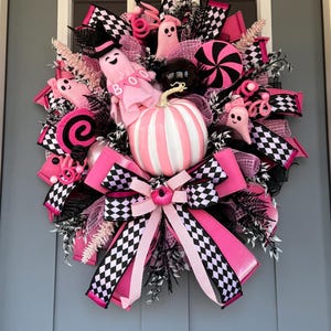 Op de afbeelding: Een decoratieve krans met een centrale wit en roze gestreepte pompoen, omringd door roze spookfiguren met de tekst BOO, zwart-wit geblokte linten, roze gaas en zwarte bladeren. Het ontwerp bevat ronde, snoepachtige decoraties.