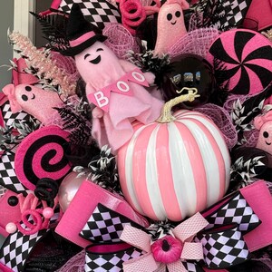 Op de afbeelding: Een decoratieve krans met een centrale roze en wit gestreepte pompoen, roze spookfiguren, waarvan er &eacute;&eacute;n een zwarte heksenhoed draagt en een spandoek met de tekst BOO vasthoudt, zwart-wit geblokte linten, roze mesh-accenten en zwarte ornamenten.