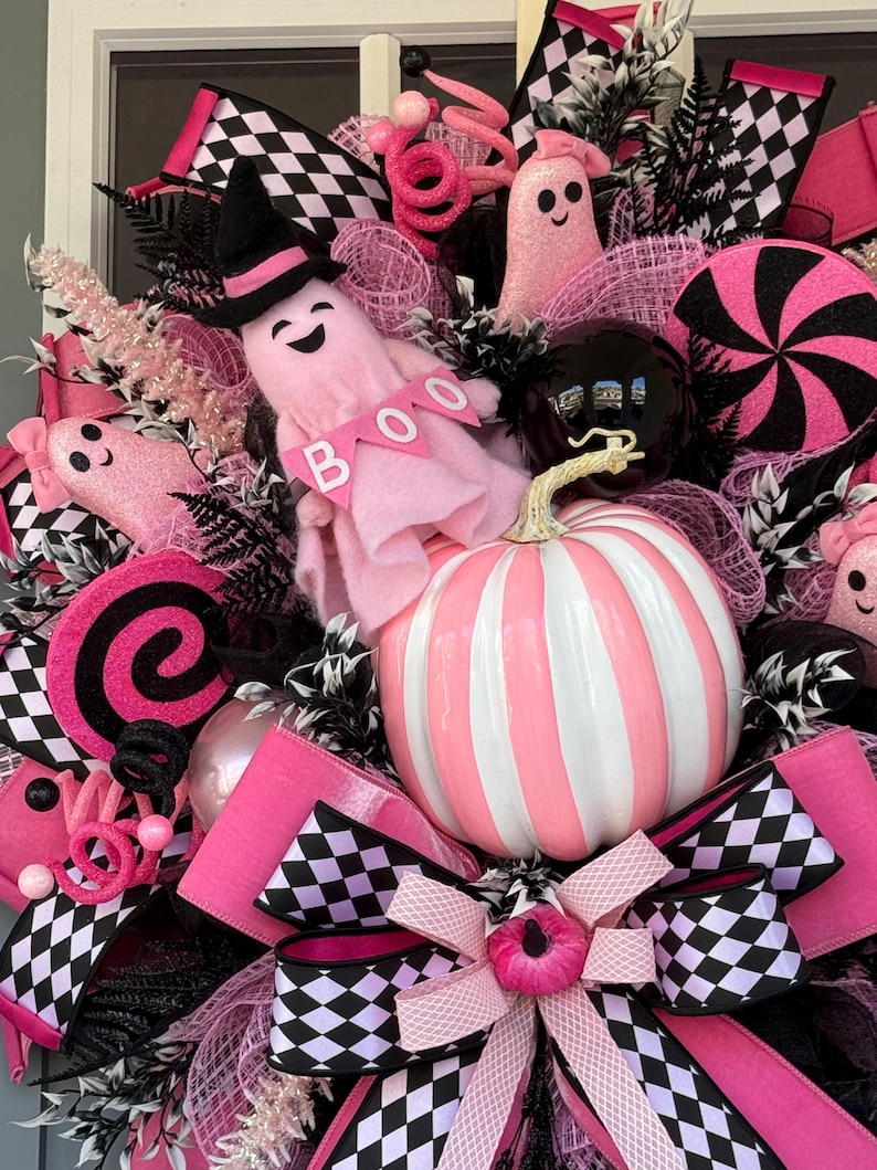 Op de afbeelding: Een decoratieve krans met een centrale roze en wit gestreepte pompoen, roze spookfiguren, waarvan er &eacute;&eacute;n een zwarte heksenhoed draagt en een spandoek met de tekst BOO vasthoudt, zwart-wit geblokte linten, roze mesh-accenten en zwarte ornamenten.