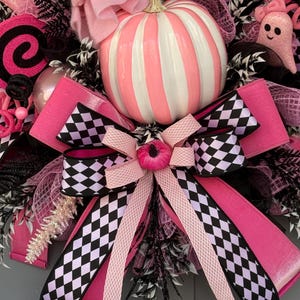 Op de afbeelding: Een decoratieve krans met een centrale gestreepte roze en witte pompoen, een grote strik met een zwart-wit geblokt patroon, roze spookfiguren met de tekst BOO, zwarte mesh-accenten en glinsterende roze en zwarte decoratieve elementen.