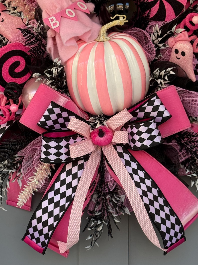 Op de afbeelding: Een decoratieve krans met een centrale gestreepte roze en witte pompoen, een grote strik met een zwart-wit geblokt patroon, roze spookfiguren met de tekst BOO, zwarte mesh-accenten en glinsterende roze en zwarte decoratieve elementen.