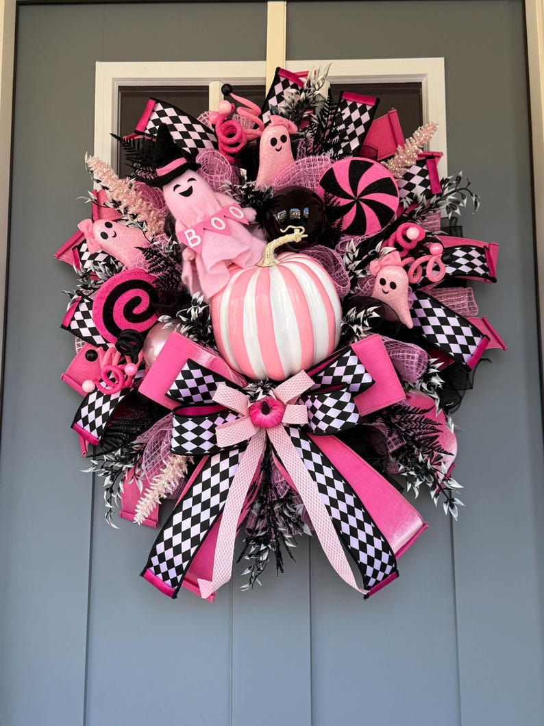 Op de afbeelding: Een decoratieve krans met een centrale wit en roze gestreepte pompoen, omringd door roze spookfiguren met de tekst BOO, zwart-wit geblokte linten, roze gaas en zwarte bladeren. Het ontwerp bevat ronde, snoepachtige decoraties.
