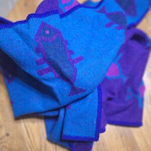 May include: A folded blue and purple blanket with a fish and geometric pattern. The blanket has a blue background with purple fish and geometric shapes. The edges are trimmed with a dark blue stitching.