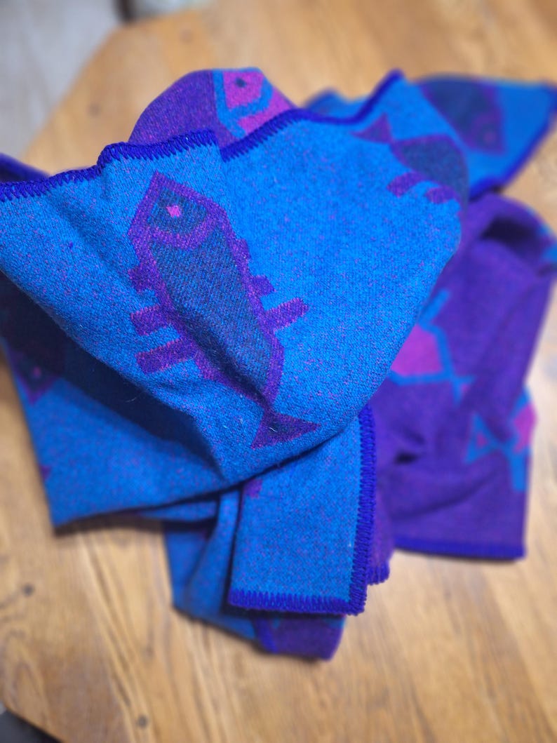May include: A folded blue and purple blanket with a fish and geometric pattern. The blanket has a blue background with purple fish and geometric shapes. The edges are trimmed with a dark blue stitching.