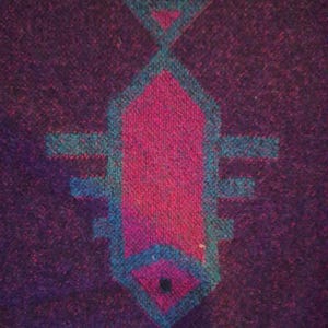 May include: A close-up of a woven textile featuring a geometric design. The central motif is a stylized fish shape in magenta and teal, set against a deep purple background. The edges of the textile have a dark border.
