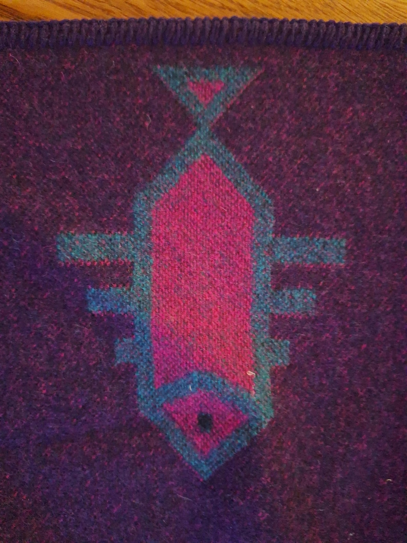 May include: A close-up of a woven textile featuring a geometric design. The central motif is a stylized fish shape in magenta and teal, set against a deep purple background. The edges of the textile have a dark border.