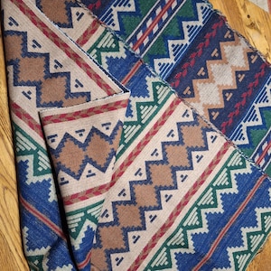 Peut inclure: Textile pli&eacute; avec un motif g&eacute;om&eacute;trique dans les tons de marron, bleu, vert et rouge. Le motif pr&eacute;sente des zigzags et des losanges. Le tissu semble &ecirc;tre une mati&egrave;re tiss&eacute;e, peut-&ecirc;tre une couverture ou un jet&eacute;.