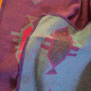 May include: A folded blanket with a geometric pattern. The blanket has a deep purple base with teal and magenta geometric shapes. The edges are finished with a purple stitched border.