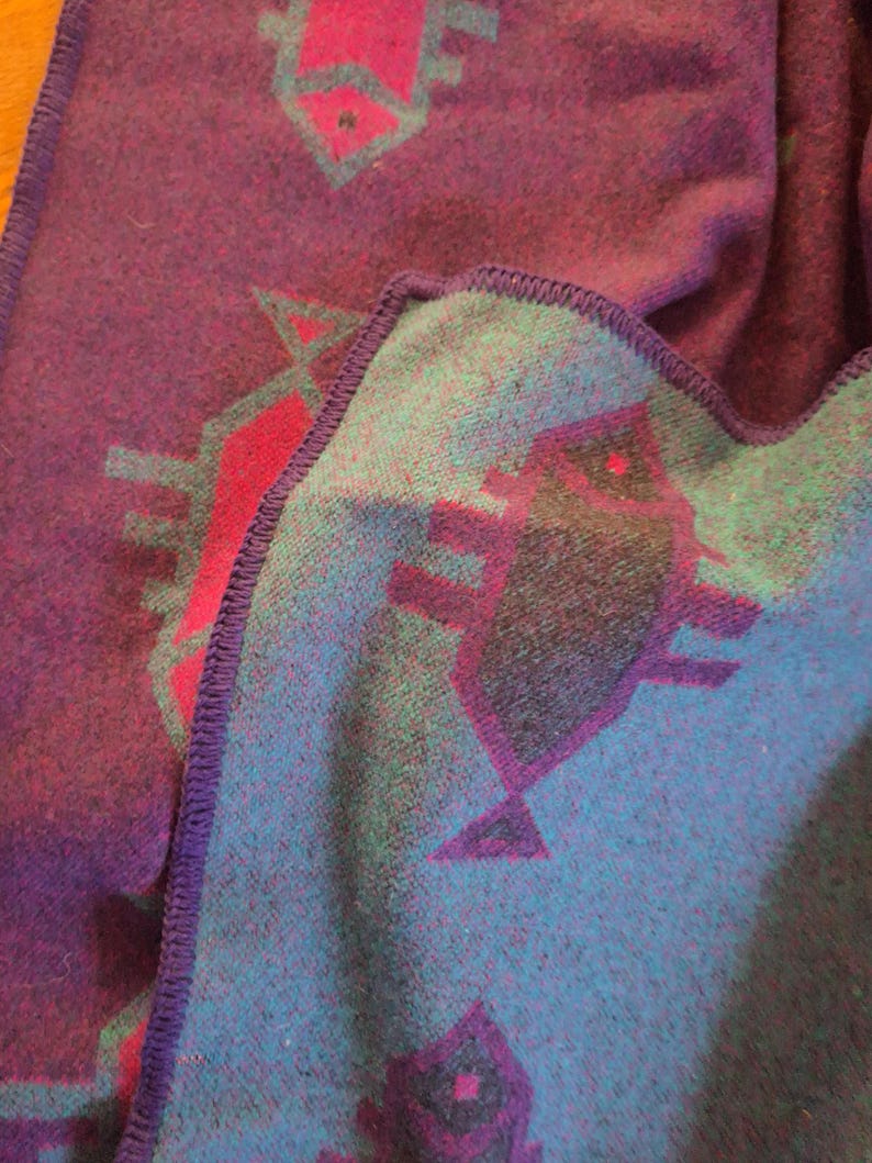 May include: A folded blanket with a geometric pattern. The blanket has a deep purple base with teal and magenta geometric shapes. The edges are finished with a purple stitched border.